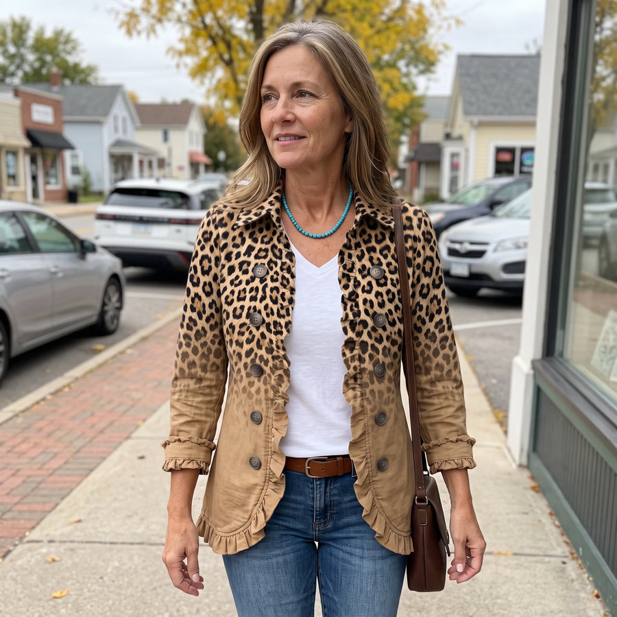 A woman stands on a sidewalk wearing a tan and brown leopard-print button-up shirt jacket over a white V-neck top, paired with blue jeans and a brown shoulder bag.
